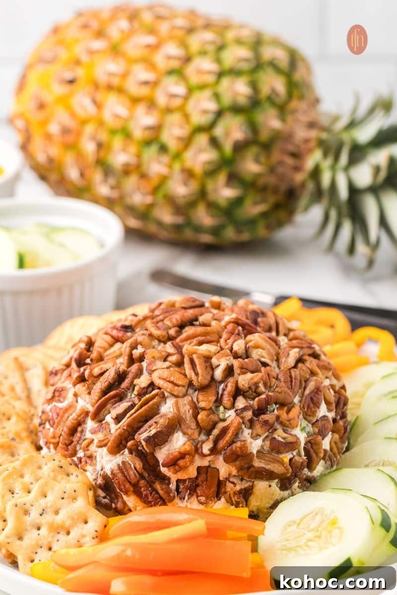 The pecan-crusted cheese ball appetizer served on a platter with sliced cucumbers and orange bell peppers in the foreground, with a whole fresh pineapple sitting on the counter in the background.
