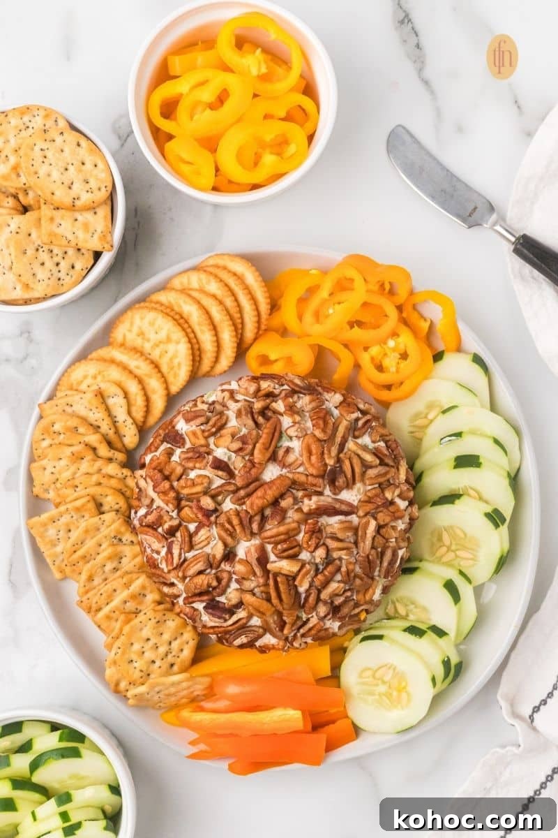 Overhead view of a completed pecan cheese ball appetizer on a platter, surrounded by circular rows of round crackers, sliced cucumbers, and yellow and orange bell pepper rings.