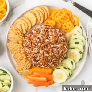 A round cream cheese ball coated entirely in pecans, served on a white platter with crackers, sliced cucumbers, and rings of yellow and orange bell peppers.