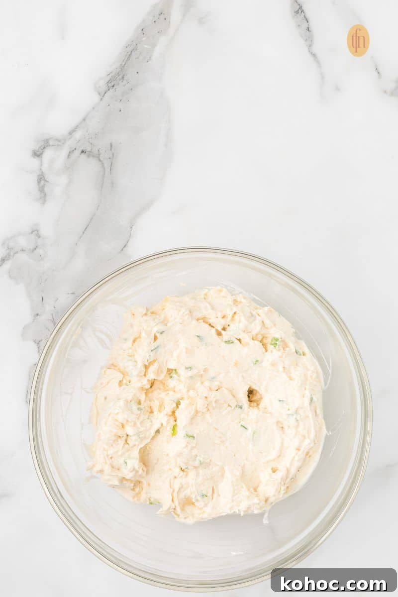 A clear glass bowl holding the fully mixed cheese ball ingredients—a creamy, off-white mixture with visible green pieces of seasoning or pepper.