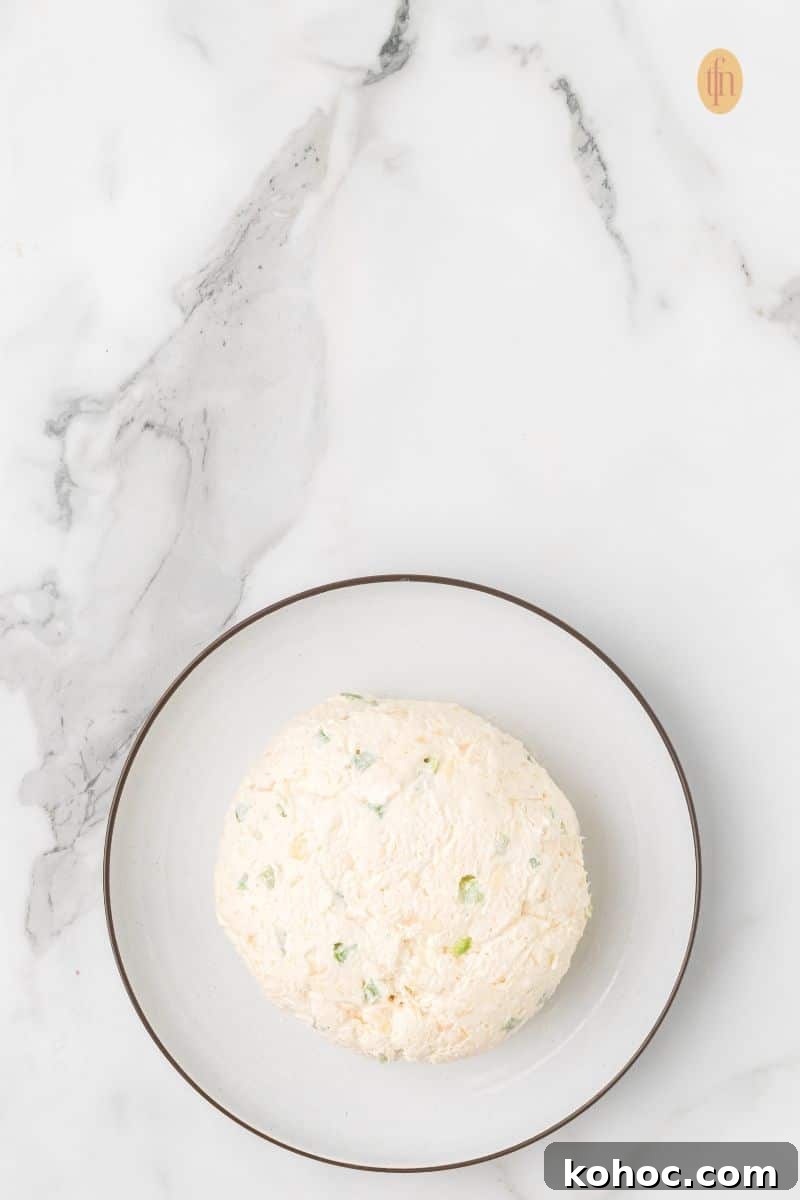 Top-down view of a round, smooth cream cheese ball (without a pecan coating) resting on a small white plate with a brown rim, set against a marble background.