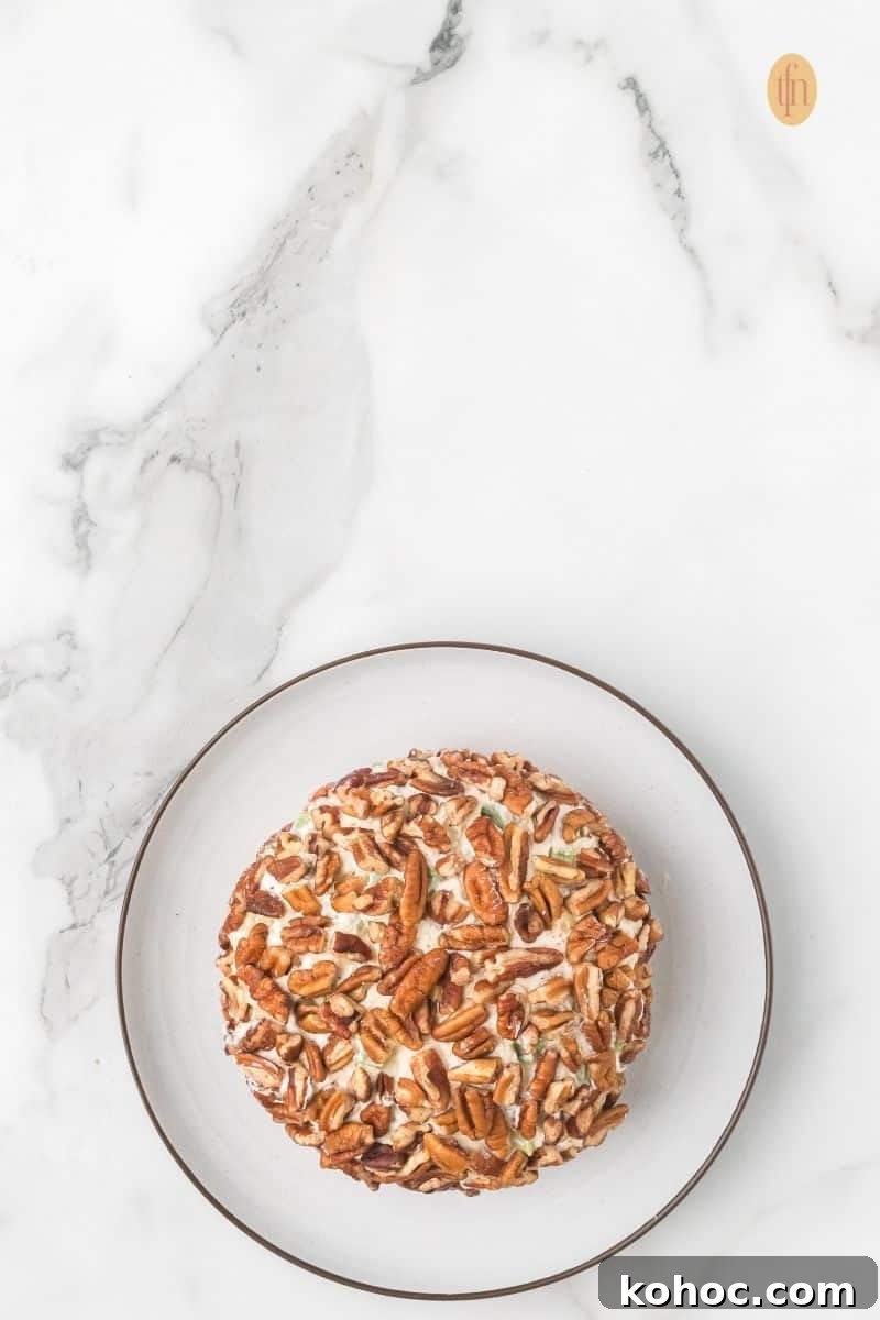 Top-down view of a cheese ball that has just been coated entirely with pecan halves, resting on a small white plate against a marble countertop.