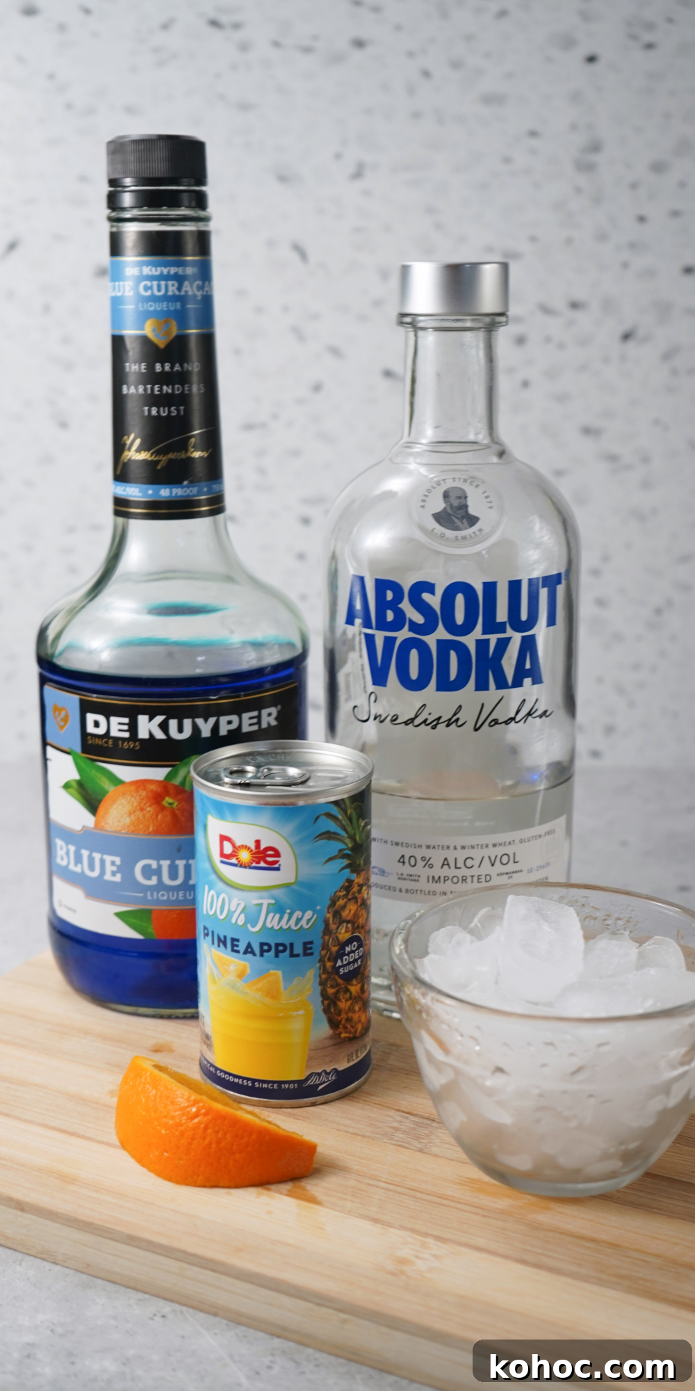 Ingredients for an ocean water cocktail arranged on a wooden board, including De Kuyper Blue Curaçao, Absolut Vodka, Dole 100% Pineapple Juice, a bowl of ice, and an orange slice.