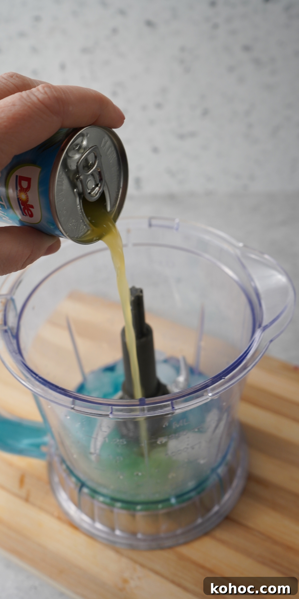 Pineapple juice being poured from a can into a blender containing ice and blue liquid, presumably Blue Curaçao and vodka, for making an ocean water cocktail.