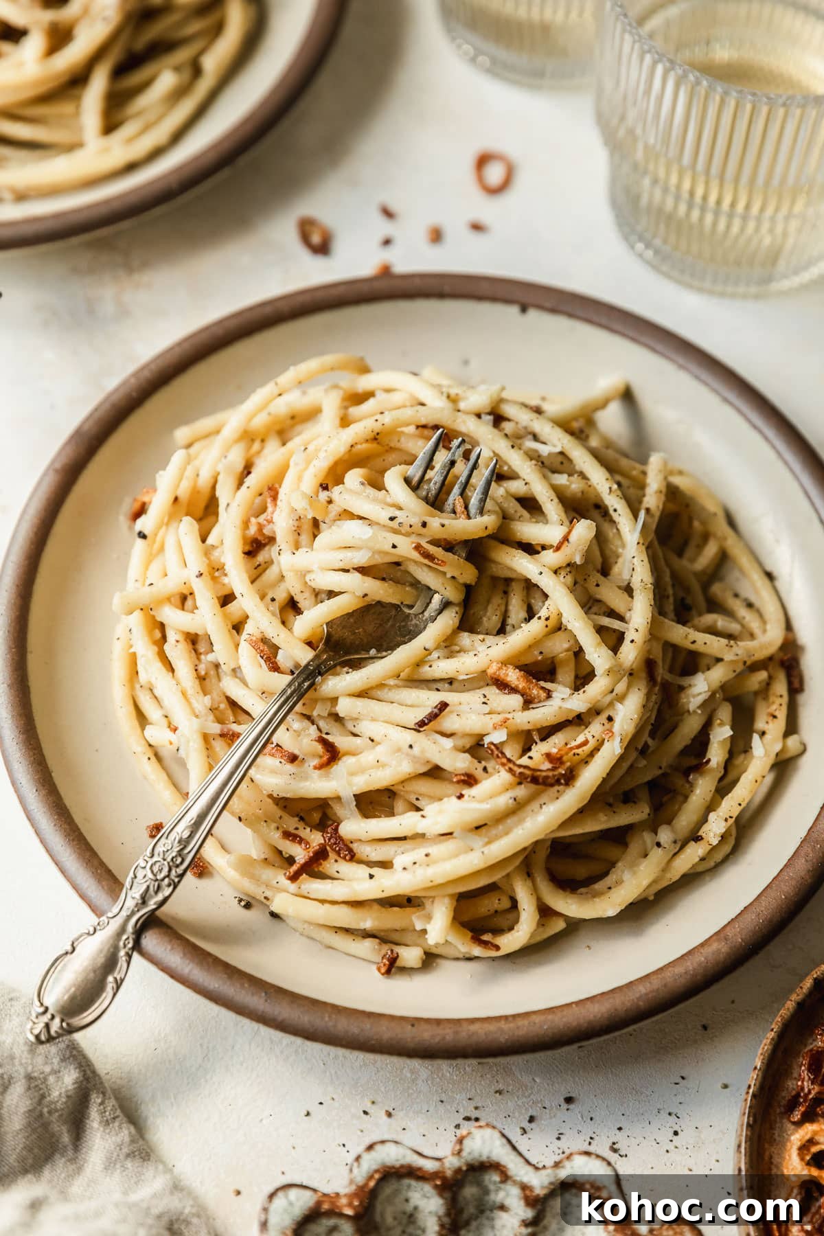 Crispy Shallot Bucatini Cacio e Pepe Recipe 8 A stoneware plate of bucatini cacio e pepe with fried shallots on a tan counter next to glasses of white wine and a brown bowl of fried shallots.