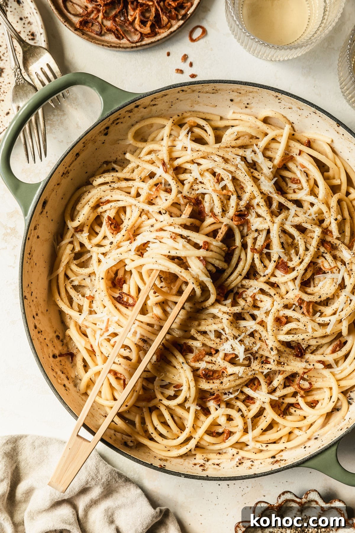 Crispy Shallot Bucatini Cacio e Pepe Recipe 9 A green and white pan with bucatini cacio e pepe on a tan counter next to white wine and brown bowls of pepper and fried shallots.