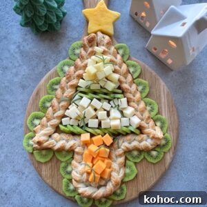 A Christmas tree-shaped pastry braid topped with cheese cubes and surrounded by kiwi slices on a wooden board, with decorative ceramic houses in the background.