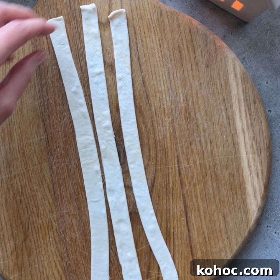 Three strips of dough are laid out side by side on a round wooden cutting board, with a hand reaching toward them from the left.
