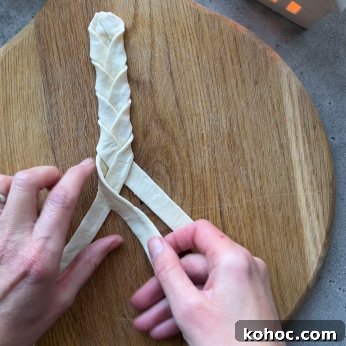 Two hands braiding three strips of dough on a round wooden board.