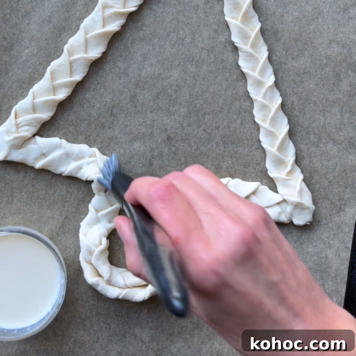 A hand uses a pastry brush to apply cream to braided dough shaped like a tree on parchment paper.