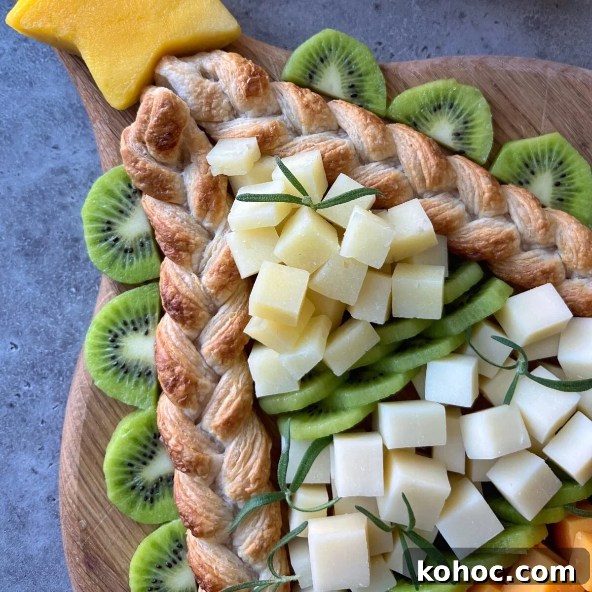 A cheese board is arranged decoratively with sliced kiwi, cubed white cheese, braided puff pastry, and a star-shaped cheese piece on top.