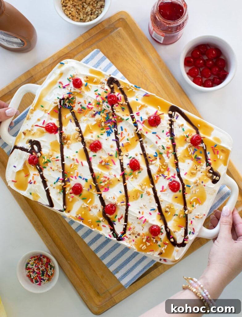 Banana Split Ice Cream Sandwich Cake Recipe for Summer Parties 2 A large ice cream sundae dessert in a white dish, topped with whipped cream, rainbow sprinkles, maraschino cherries, chocolate syrup, and caramel sauce. Surrounding bowls hold cherries, sprinkles, and syrup.