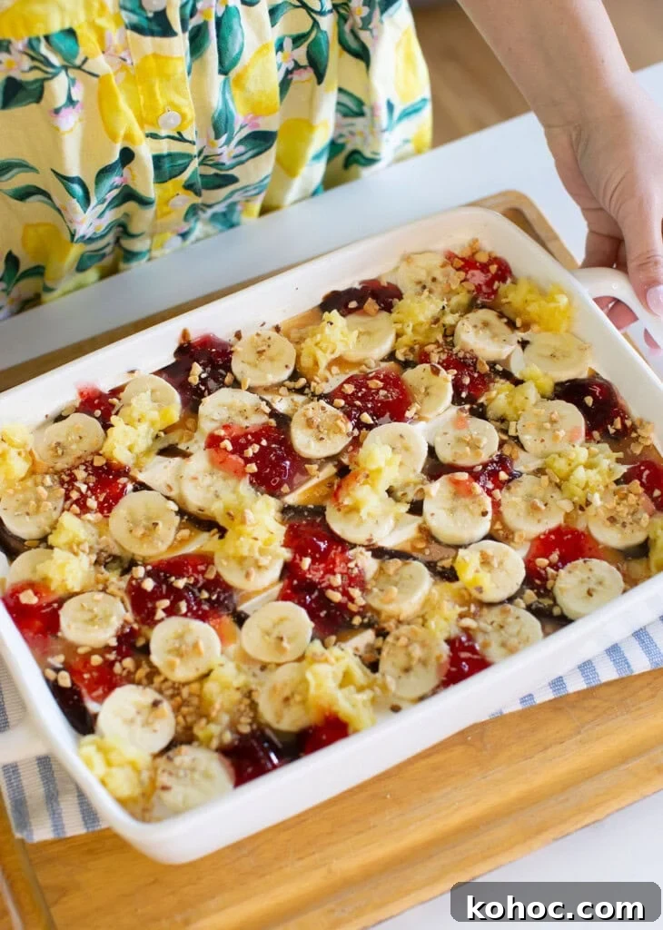Banana Split Ice Cream Sandwich Cake Recipe for Summer Parties 11 A person in a yellow, lemon-patterned dress holds a white baking dish filled with sliced bananas, dollops of red jelly, crushed nuts, and other toppings, placed on a wooden tray.