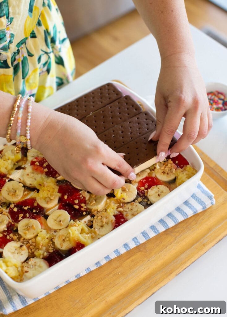 Banana Split Ice Cream Sandwich Cake Recipe for Summer Parties 12 A person places chocolate ice cream sandwiches on top of a dessert layered with banana slices, pineapple, strawberry sauce, and crushed nuts in a white dish. The dish sits on a striped cloth on a wooden tray.