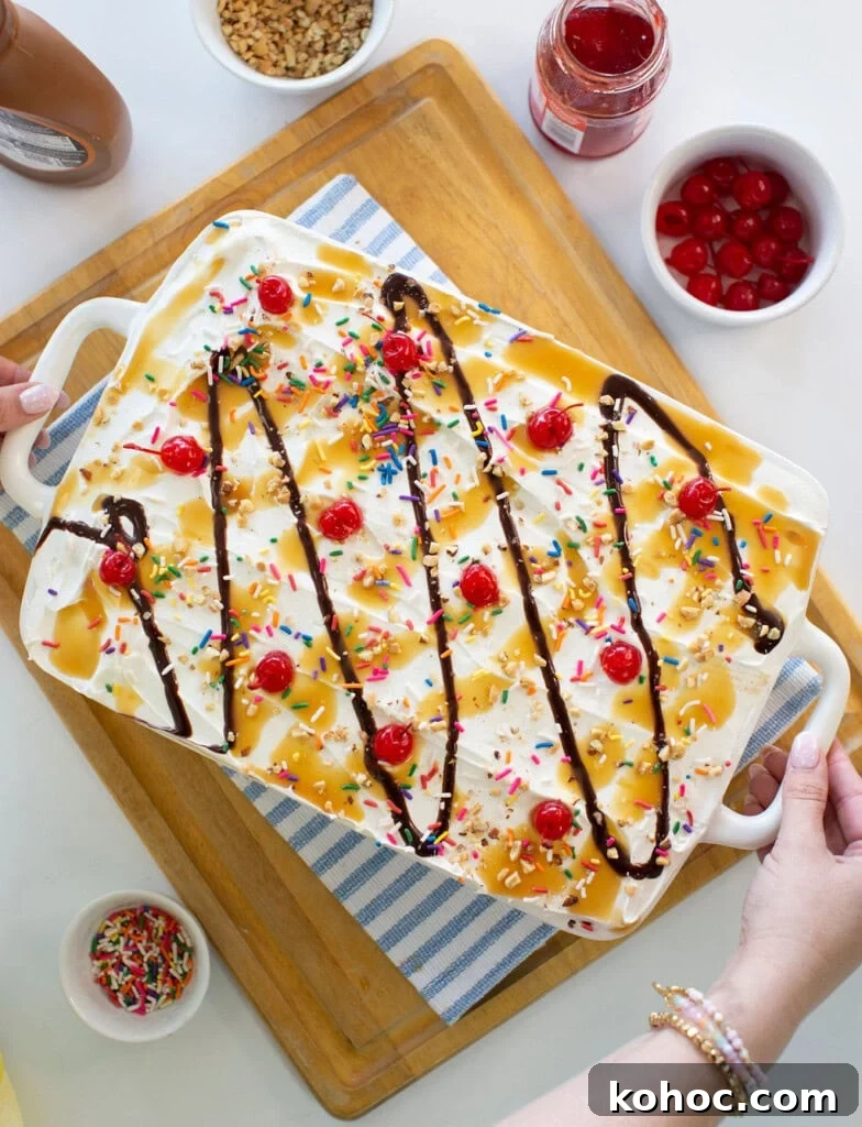 Banana Split Ice Cream Sandwich Cake Recipe for Summer Parties 13 A large ice cream sundae dessert in a white dish, topped with whipped cream, rainbow sprinkles, maraschino cherries, chocolate syrup, and caramel sauce. Surrounding bowls hold cherries, sprinkles, and syrup.