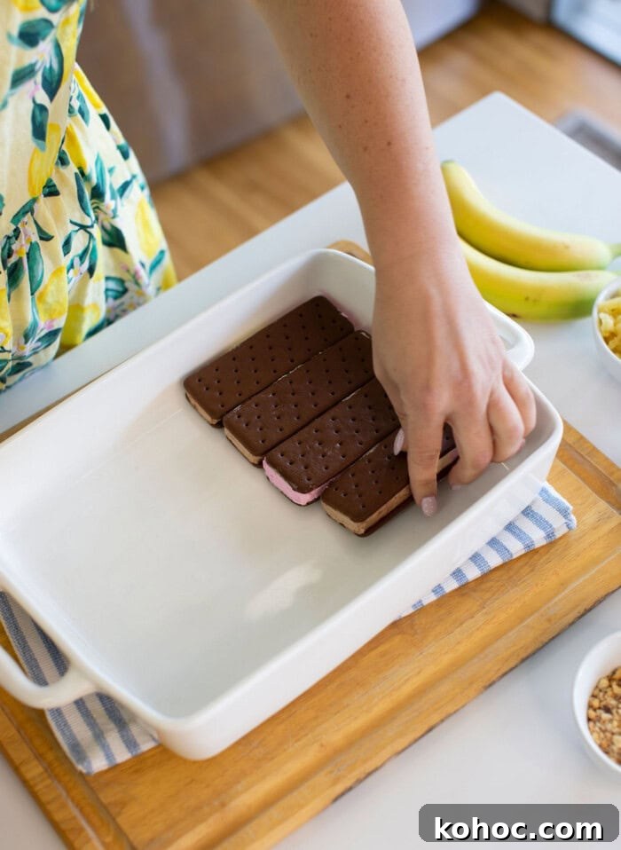 Banana Split Ice Cream Sandwich Cake Recipe for Summer Parties 15 A person places ice cream sandwiches in a white baking dish on a wooden board; bananas and small bowls with toppings are nearby.