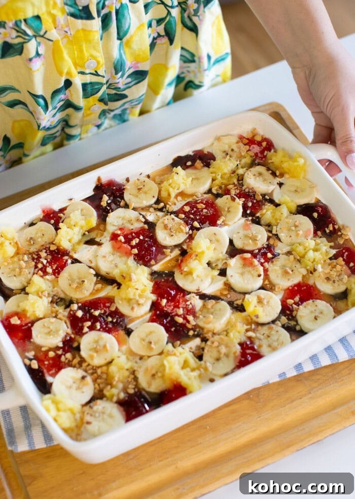 Banana Split Ice Cream Sandwich Cake Recipe for Summer Parties 16 A person in a yellow, lemon-patterned dress holds a white baking dish filled with sliced bananas, dollops of red jelly, crushed nuts, and other toppings, placed on a wooden tray.