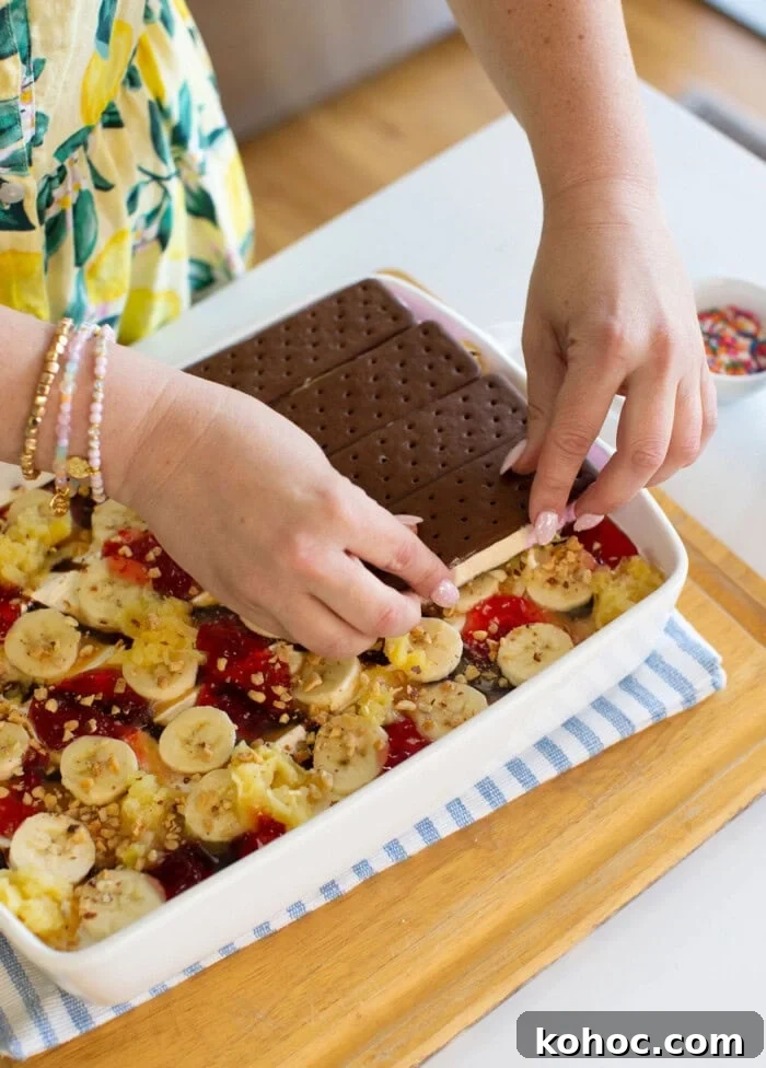 Banana Split Ice Cream Sandwich Cake Recipe for Summer Parties 17 A person places chocolate ice cream sandwiches on top of a dessert layered with banana slices, pineapple, strawberry sauce, and crushed nuts in a white dish. The dish sits on a striped cloth on a wooden tray.