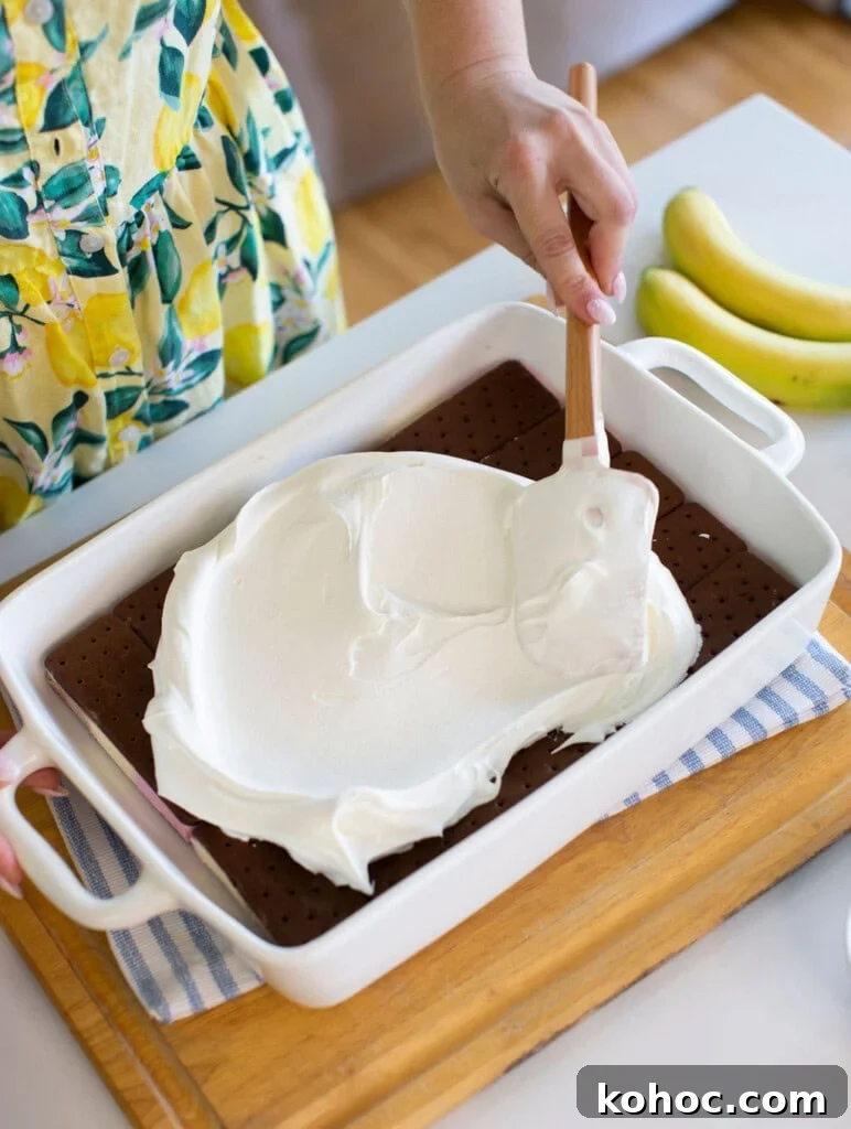 Banana Split Ice Cream Sandwich Cake Recipe for Summer Parties 7 A person spreads a thick layer of whipped topping over ice cream sandwiches in a white baking dish. Two bananas and a blue-striped cloth are on the counter nearby. The person is wearing a yellow, floral-patterned dress.