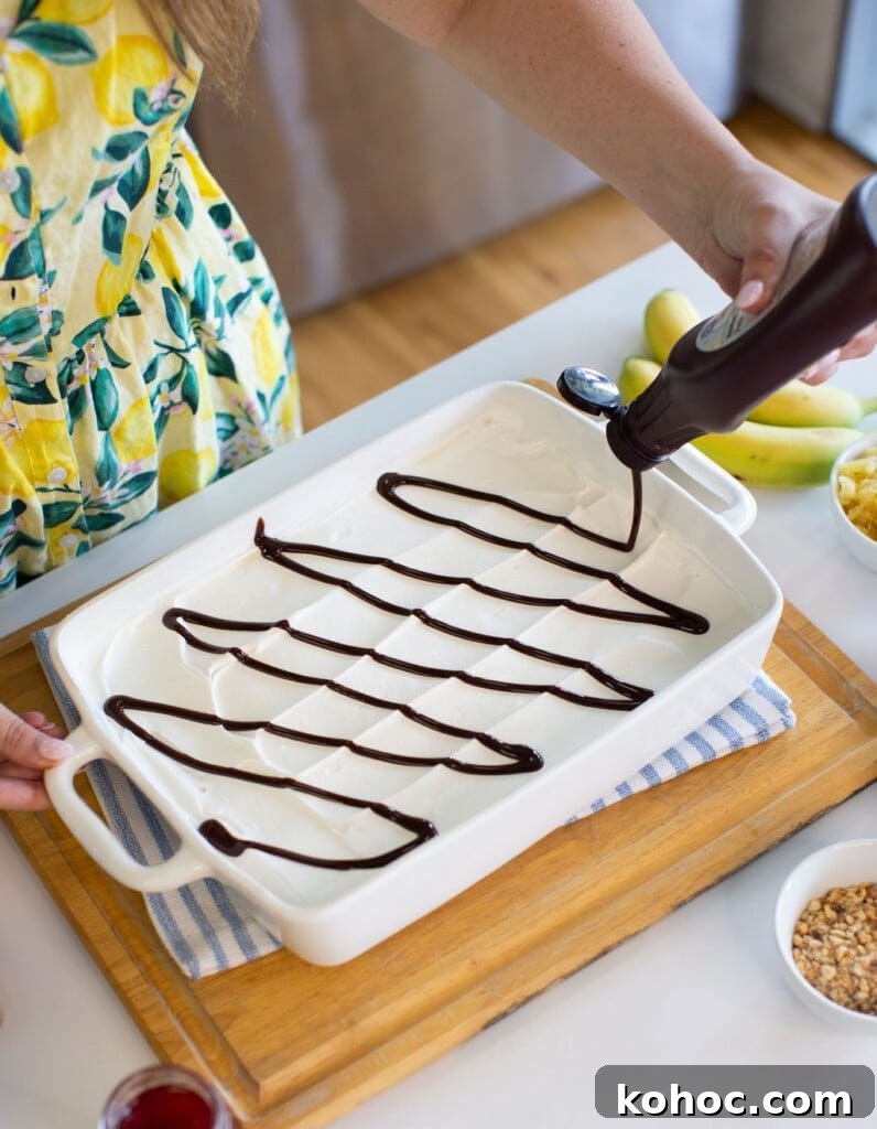 Banana Split Ice Cream Sandwich Cake Recipe for Summer Parties 8 A person in a yellow dress with a lemon pattern is drizzling chocolate syrup in a grid pattern over a layer of vanilla ice cream in a white rectangular dish on a wooden tray. Nearby are bananas and bowls with chopped nuts.