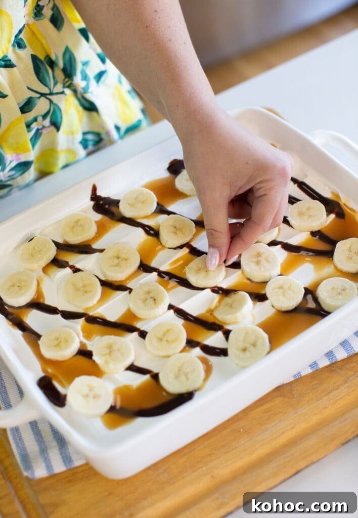 Banana Split Ice Cream Sandwich Cake Recipe for Summer Parties 9 A hand places banana slices on a white dish drizzled with caramel and chocolate sauce. The person is wearing a yellow and green dress with a lemon pattern. The dish rests on a blue-striped towel and wooden surface.