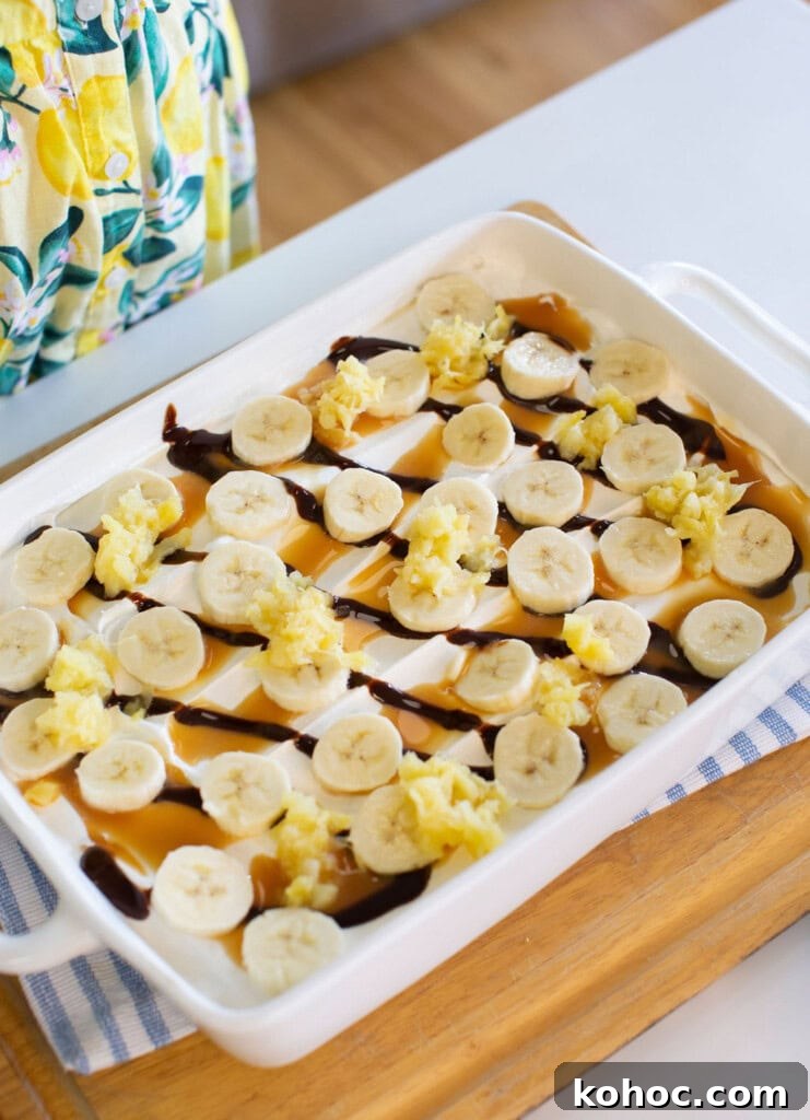 Banana Split Ice Cream Sandwich Cake Recipe for Summer Parties 10 A white baking dish filled with a dessert topped with banana slices, caramel and chocolate drizzle, and small scoops of what appears to be crushed pineapple. A person in a lemon-patterned apron stands nearby.