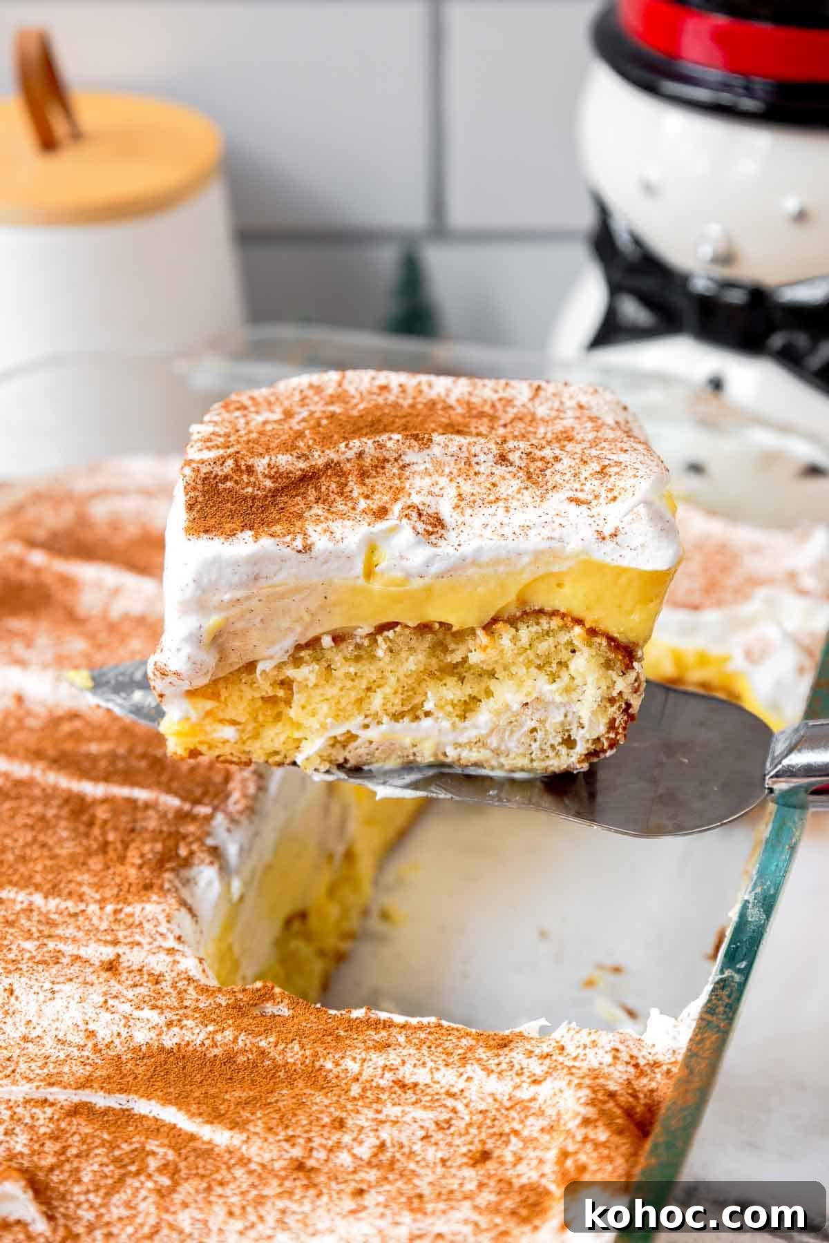 A slice of eggnog poke cake being lifted from a glass baking dish, showing layers of butter cake, eggnog pudding, and whipped topping dusted with cinnamon and nutmeg.