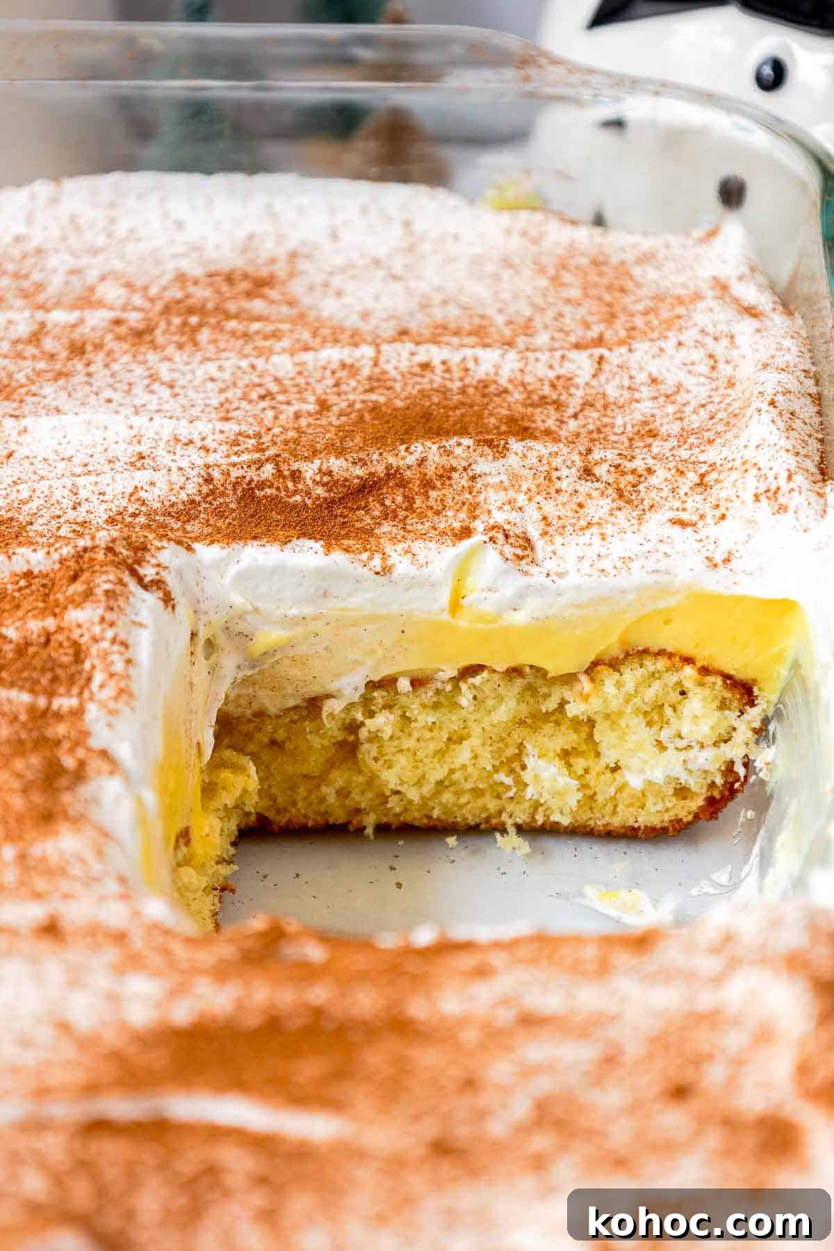 A partially sliced eggnog poke cake in a glass pan, revealing a butter cake layer and creamy eggnog pudding beneath a fluffy cinnamon-dusted whipped topping.