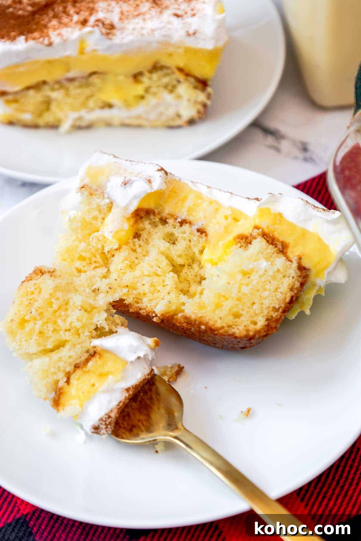 A slice of eggnog poke cake served on a white plate, featuring layers of moist butter cake, creamy eggnog pudding, and whipped topping with a dusting of cinnamon. A fork holds a bite-sized piece showing the fluffy texture and layered filling.