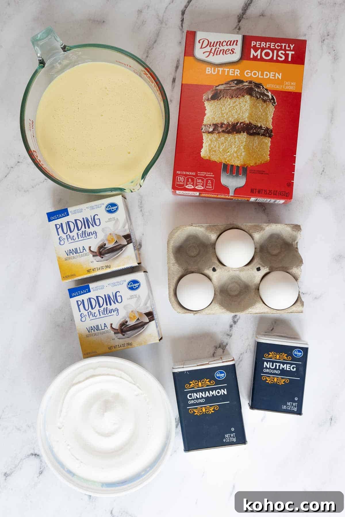 Overhead view of ingredients for an eggnog poke cake, including boxed butter cake mix, instant vanilla pudding, eggs, whipped topping, cinnamon, nutmeg, and a glass measuring cup of prepared eggnog.