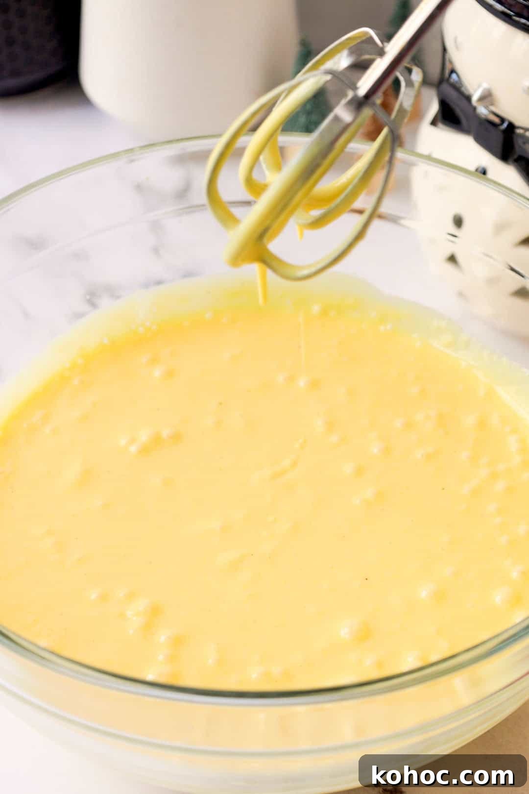 Smooth, yellow eggnog pudding filling being mixed in a bowl with beaters suspended above, showing a loose runny texture.
