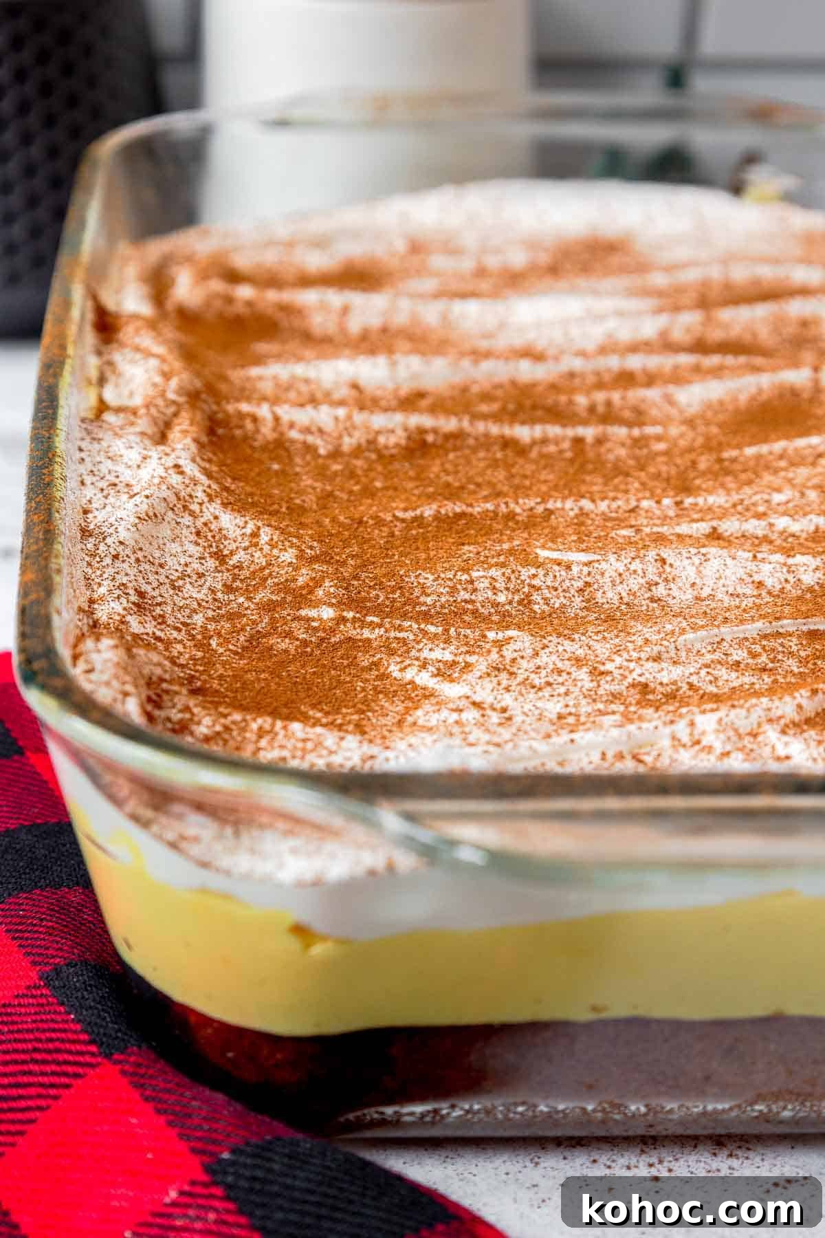 A full, uncut eggnog poke cake in a glass dish with visible layers of butter cake, eggnog filling, and whipped cream sprinkled with cinnamon.