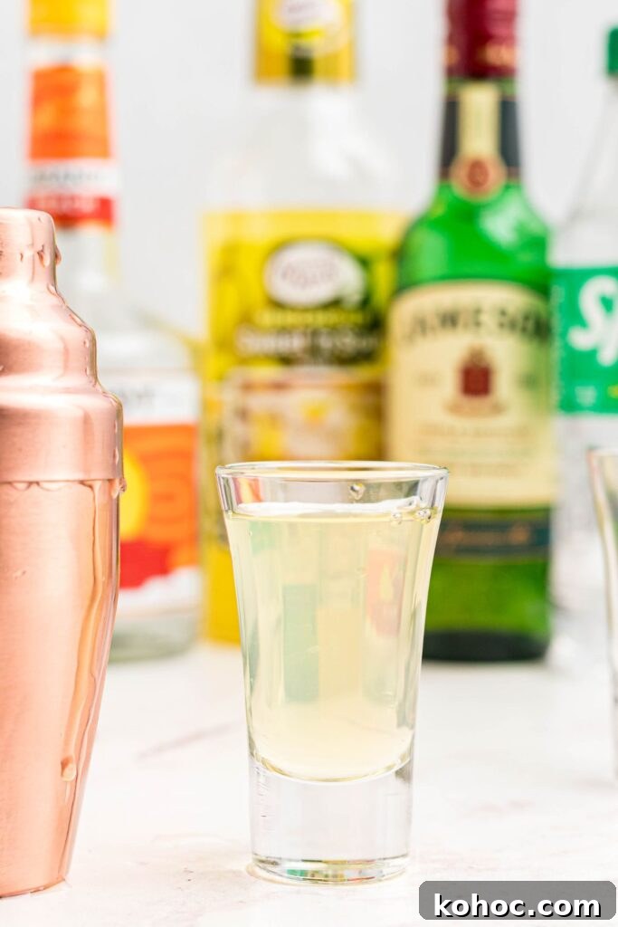 Green Tea Shot Recipe: How to Make the Classic Cocktail 2 Green tea shot in a small shote glass next to a gold cocktail shaker.