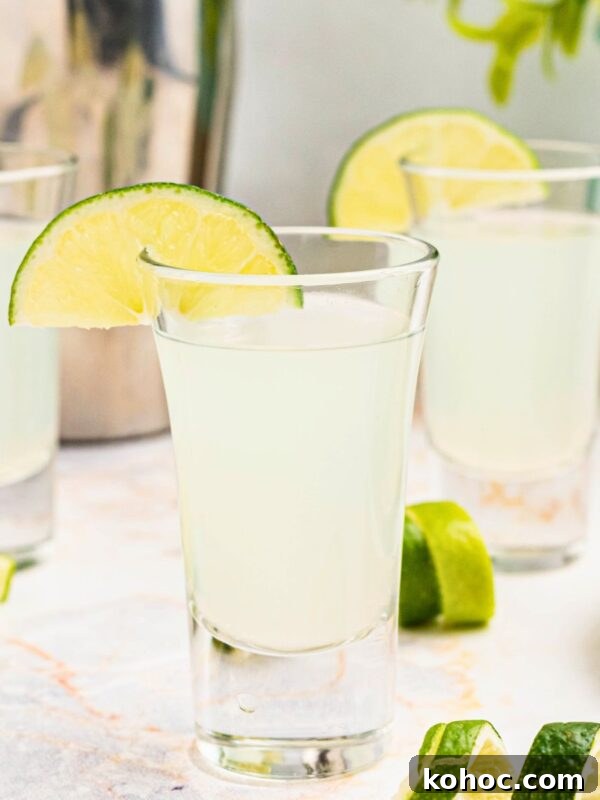 Green Tea Shot Recipe: How to Make the Classic Cocktail 7 Cold Kamikaze shots in shot glasses with lime wedges and lime curls on a marble table in front of a cocktail shaker.