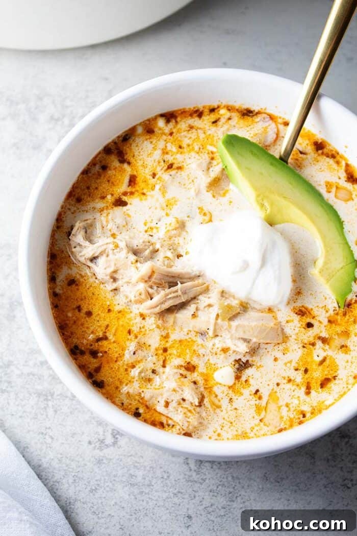 overhead view of bowl of keto white chicken chili in white serving bowl and gold serving spoon and garnished with slice of avocado and sour cream