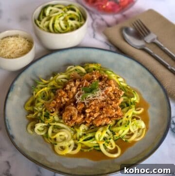 Low-Carb Chicken Bolognese Recipe for Keto Meal Prep 4 A plate of zucchini noodles topped with a keto chicken bolognese, garnished with grated cheese, sits beside bowls of more zucchini noodles and cheese on a marble surface with a napkin and cutlery.