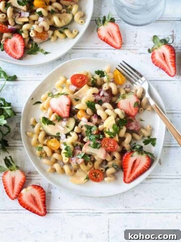 Summer pasta salad on a white plate on a wide wooden picnic table. Wooden handled fork on the side of the plate. Fresh halved strawberries on the table around the plate.