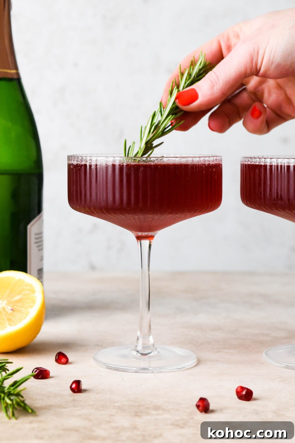 Pomegranate Champagne Cocktail with Gin or Vodka for Holidays 2 Straight on shot of a bright red bubbly festive pomegranate champagne cocktail garnished with a sprig of rosemary and sitting next to a piece of pomegranate fruit and a lemon half.