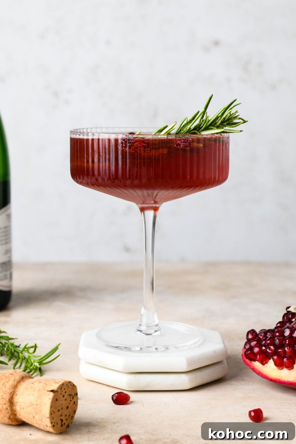 Pomegranate Champagne Cocktail with Gin or Vodka for Holidays 15 Straight on shot of a bright red bubbly festive pomegranate champagne cocktail garnished with a sprig of rosemary and sitting next to a piece of pomegranate fruit and a champagne cork.