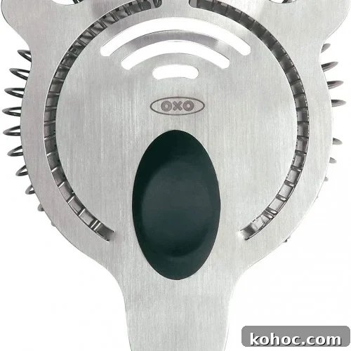Close-up of a stainless steel cocktail strainer with a black grip, made by OXO. The strainer has tightly wound metal coils and slots for draining liquid from a shaker.