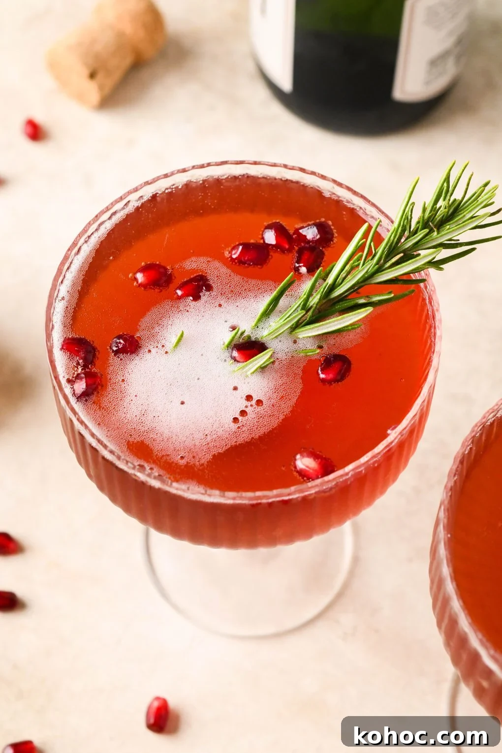 Pomegranate Champagne Cocktail with Gin or Vodka for Holidays 3 Overhead shot of festive pomegranate champagne cocktail garnished with pomegranate seeds and a sprig of rosemary. On a light brown background next to another cocktail and scattered pomegranate seeds.