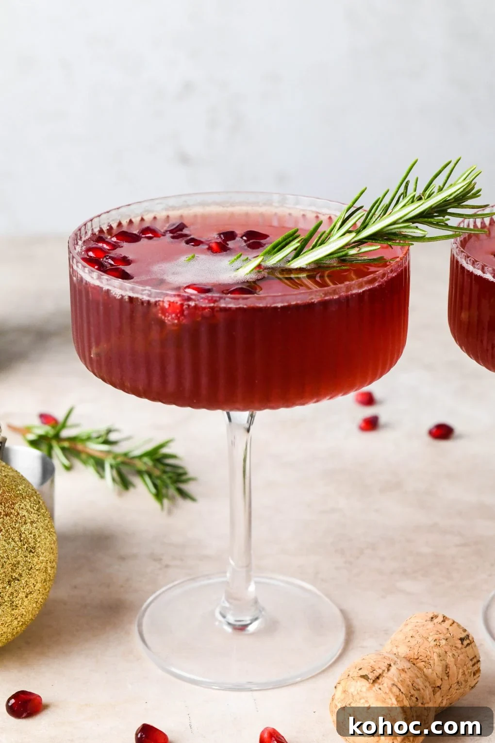 Pomegranate Champagne Cocktail with Gin or Vodka for Holidays 4 Straight on shot of a bright red bubbly festive pomegranate champagne cocktail garnished with a sprig of rosemary and sitting next to a piece of pomegranate fruit and a champagne cork.