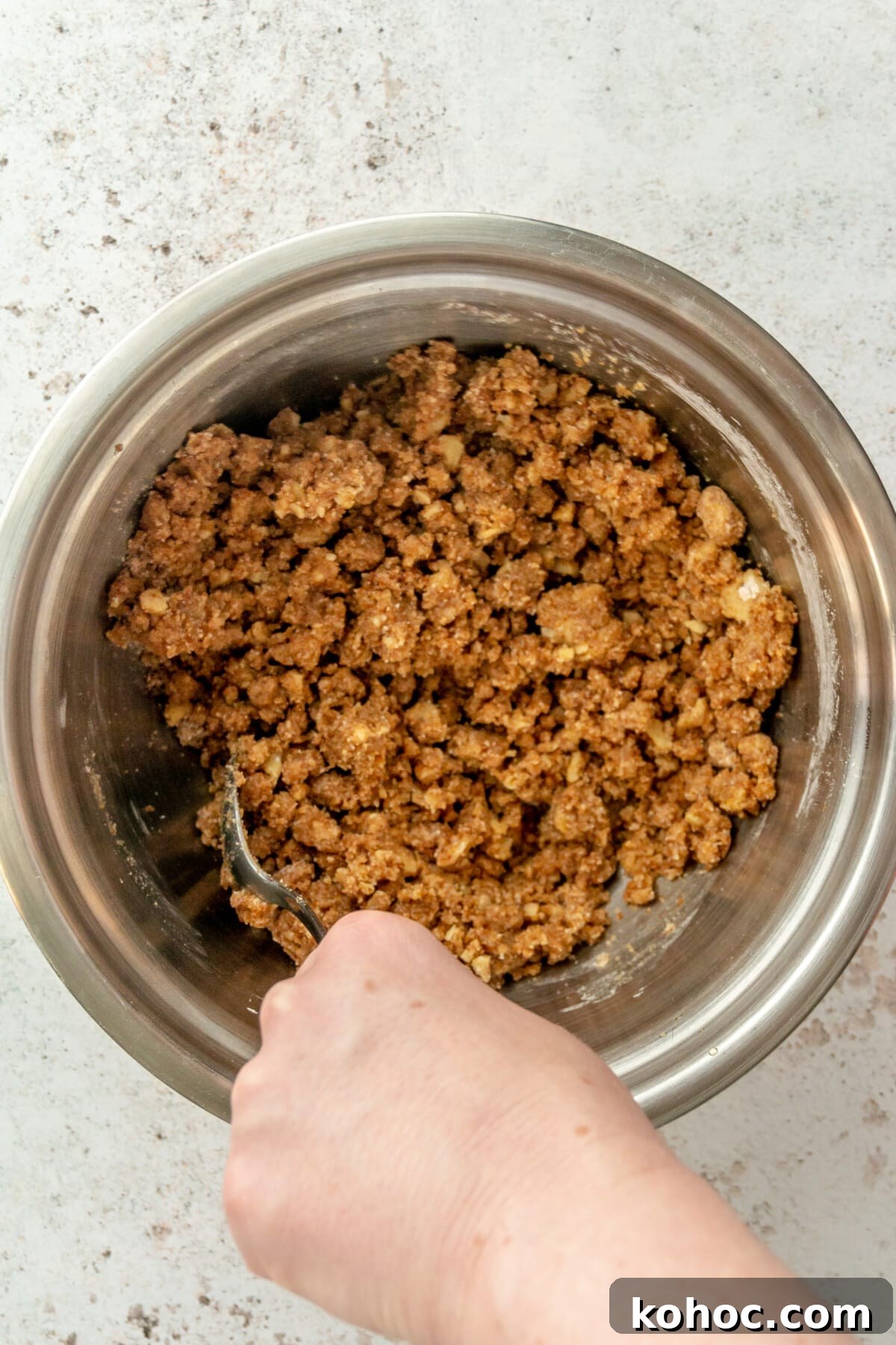 Warm Paleo Apple Crisp with Almond Flour Topping 5 A paleo crisp mixture is stirred in a stainless steel bowl on a light grey surface.