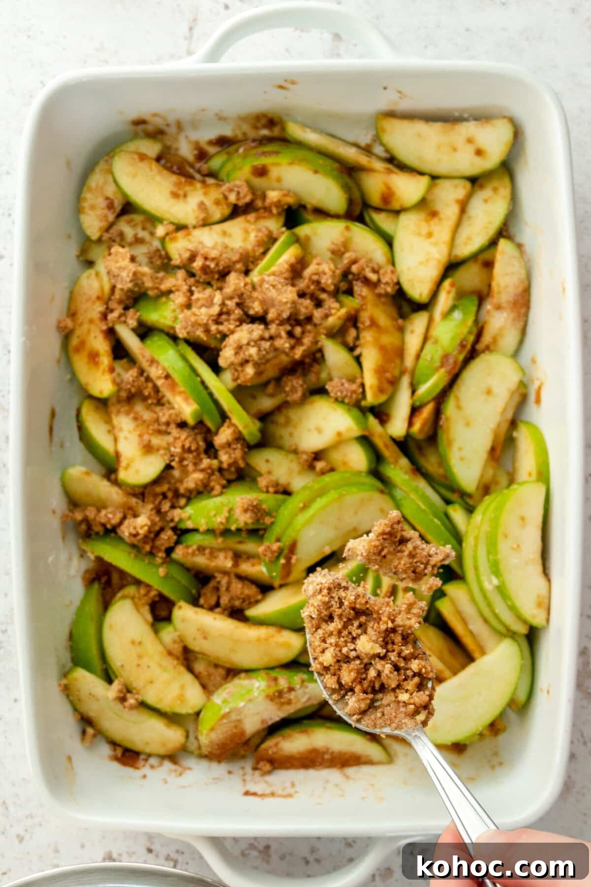 Warm Paleo Apple Crisp with Almond Flour Topping 6 Paleo crisp mixture is tossed over apples in a white ceramic rectangle dish on a light grey surface.