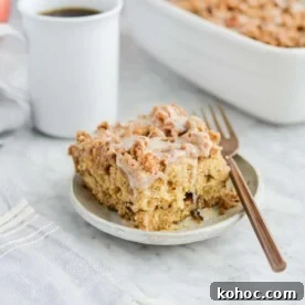 the side view of a piece of apple coffee cake on a plate sitting next to the dish of coffee cake, a mug of black coffee, and a basket of apples
