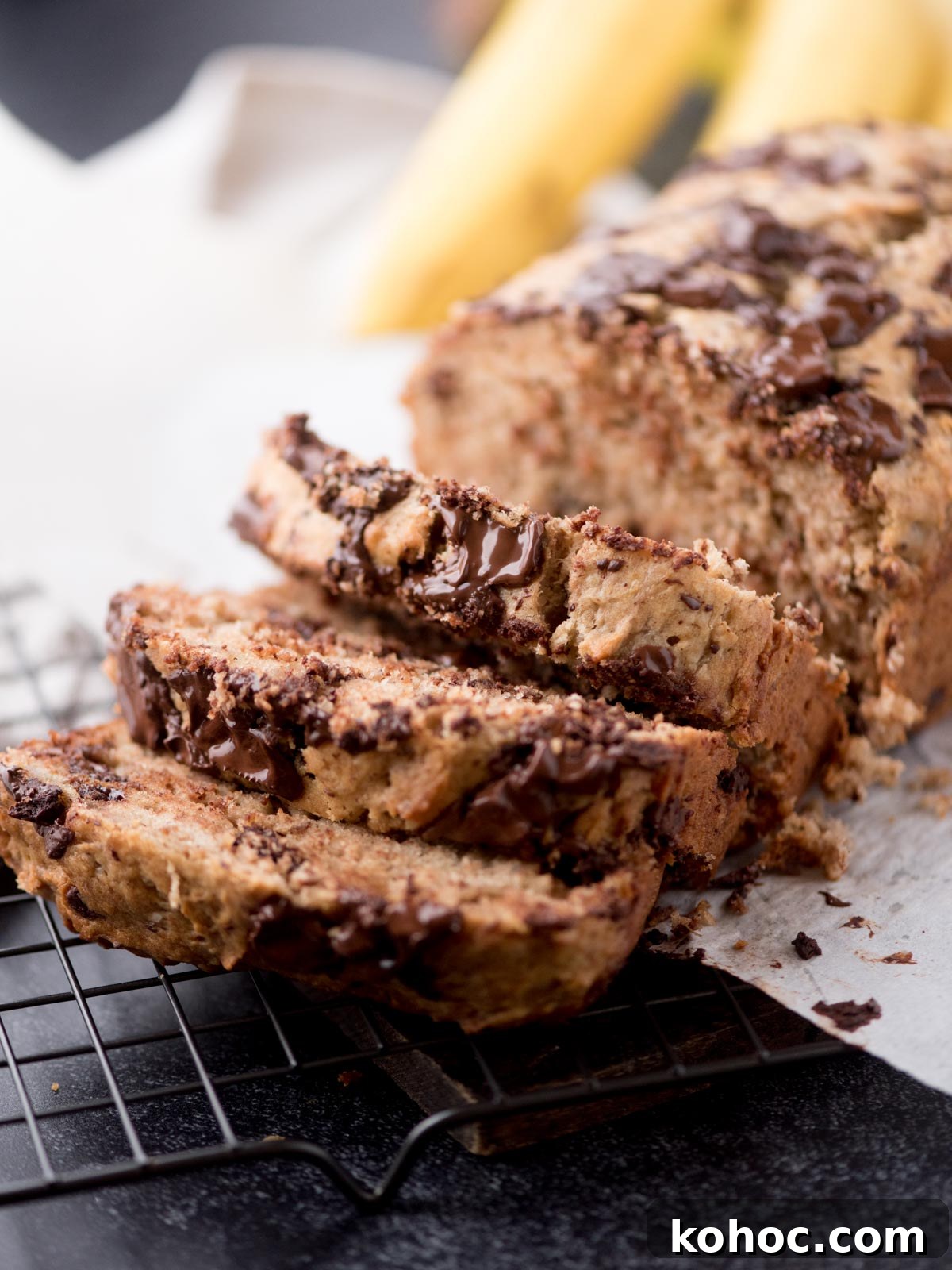Chocolate Chunk Banana Bread Recipe with Melting Chocolate Chunks 2 Sliced chocolate chip banana bread on a cooling rack, with melted chocolate chunks visible in the moist bread. Whole bananas are blurred in the background.