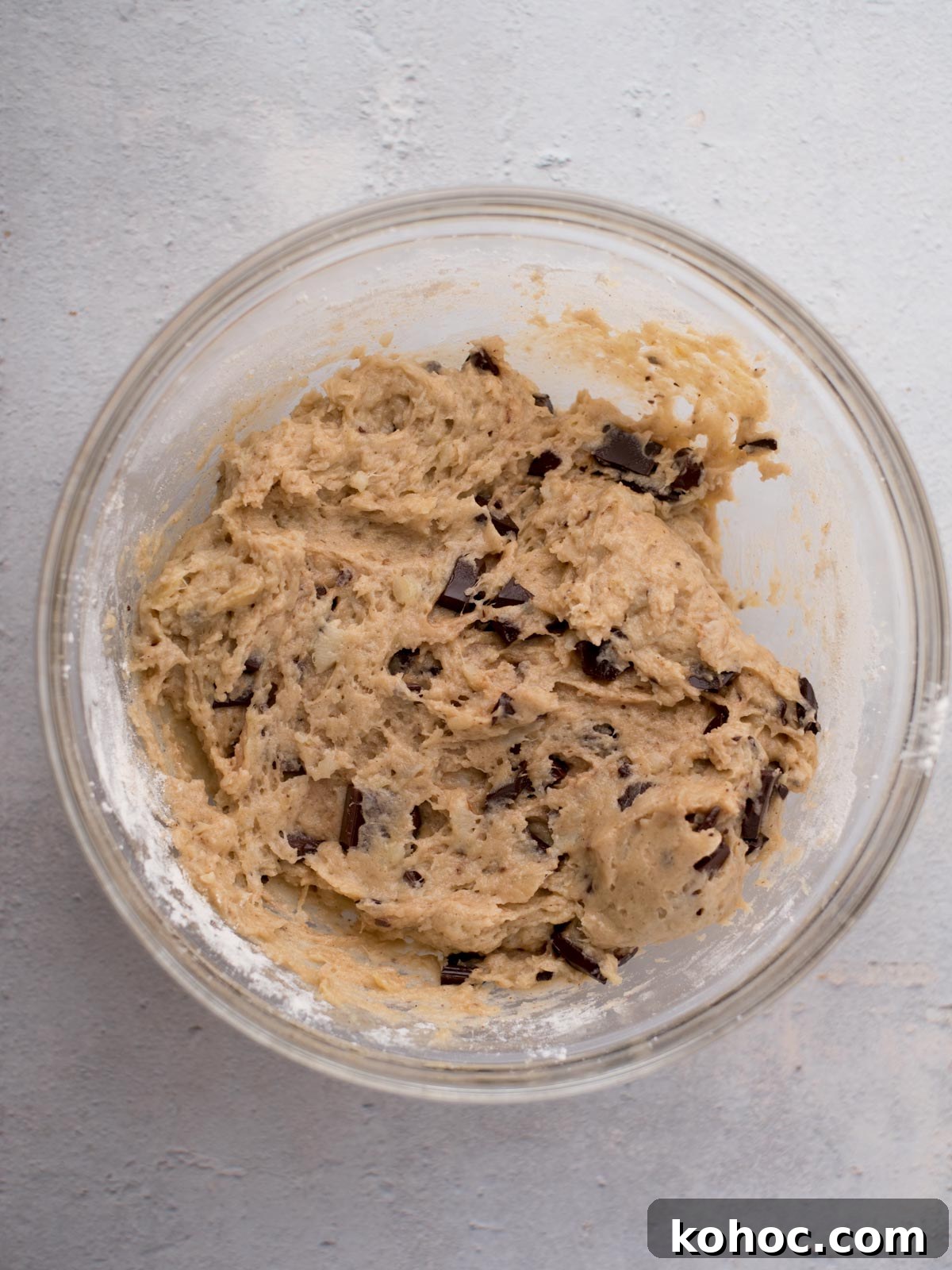 Chocolate Chunk Banana Bread Recipe with Melting Chocolate Chunks 7 A glass bowl filled with chocolate chip cookie dough, with visible chunks of chocolate, sits on a light gray surface.