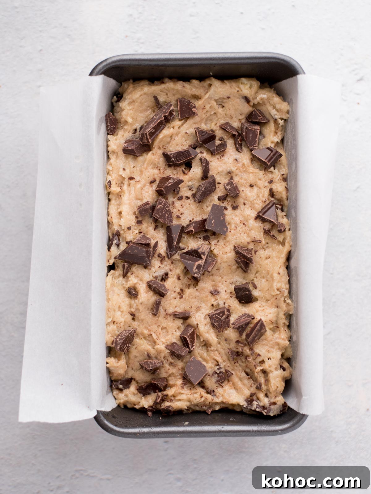 Chocolate Chunk Banana Bread Recipe with Melting Chocolate Chunks 8 A loaf pan lined with parchment paper is filled with unbaked batter topped with chunks of dark chocolate, ready to be baked. The pan sits on a light grey surface.