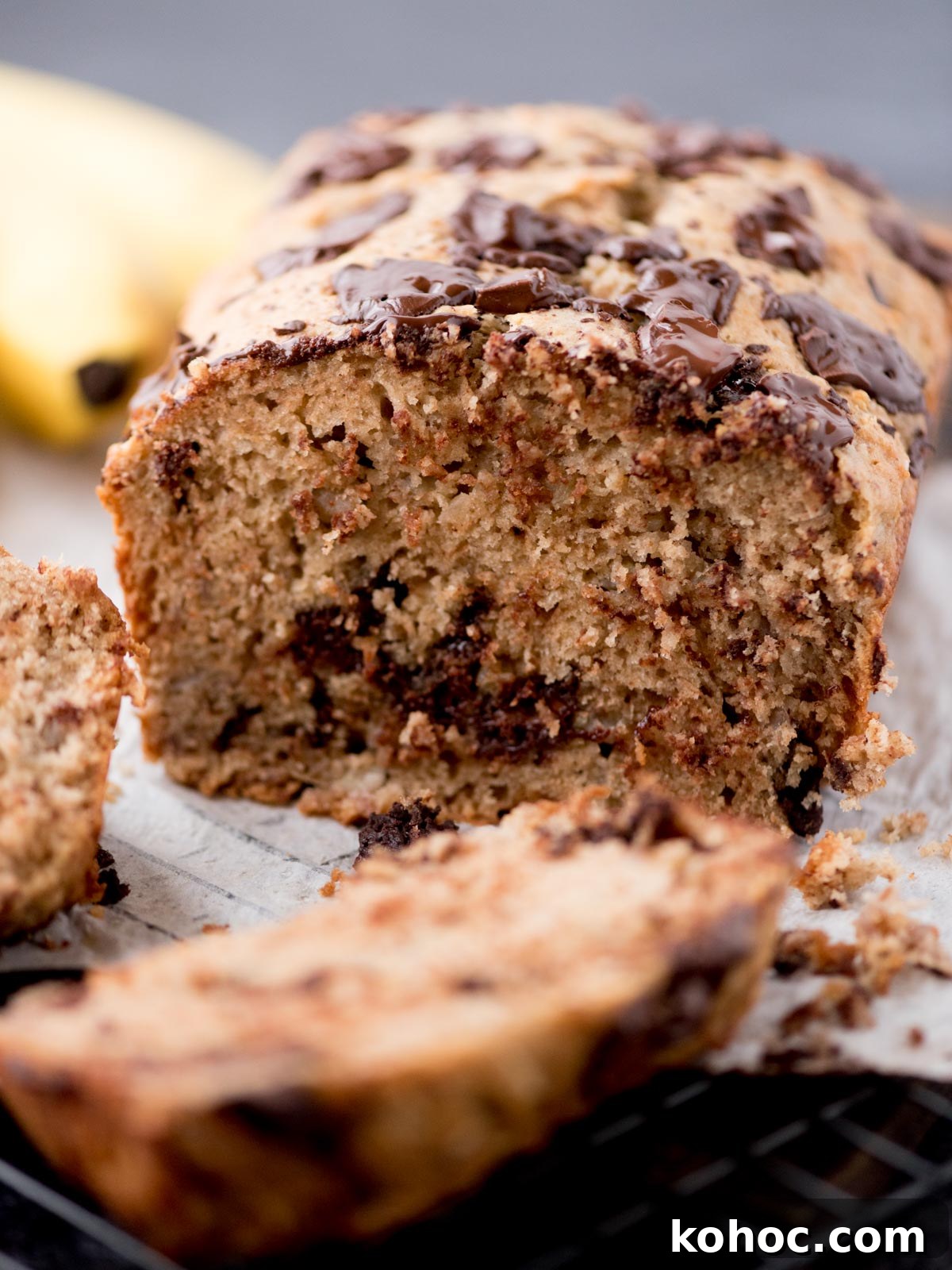Chocolate Chunk Banana Bread Recipe with Melting Chocolate Chunks 9 A close-up of a sliced loaf of banana bread with visible chocolate chunks, resting on parchment paper. The bread looks moist, with a golden-brown crust and chocolate pieces melted on top. A banana is blurred in the background.