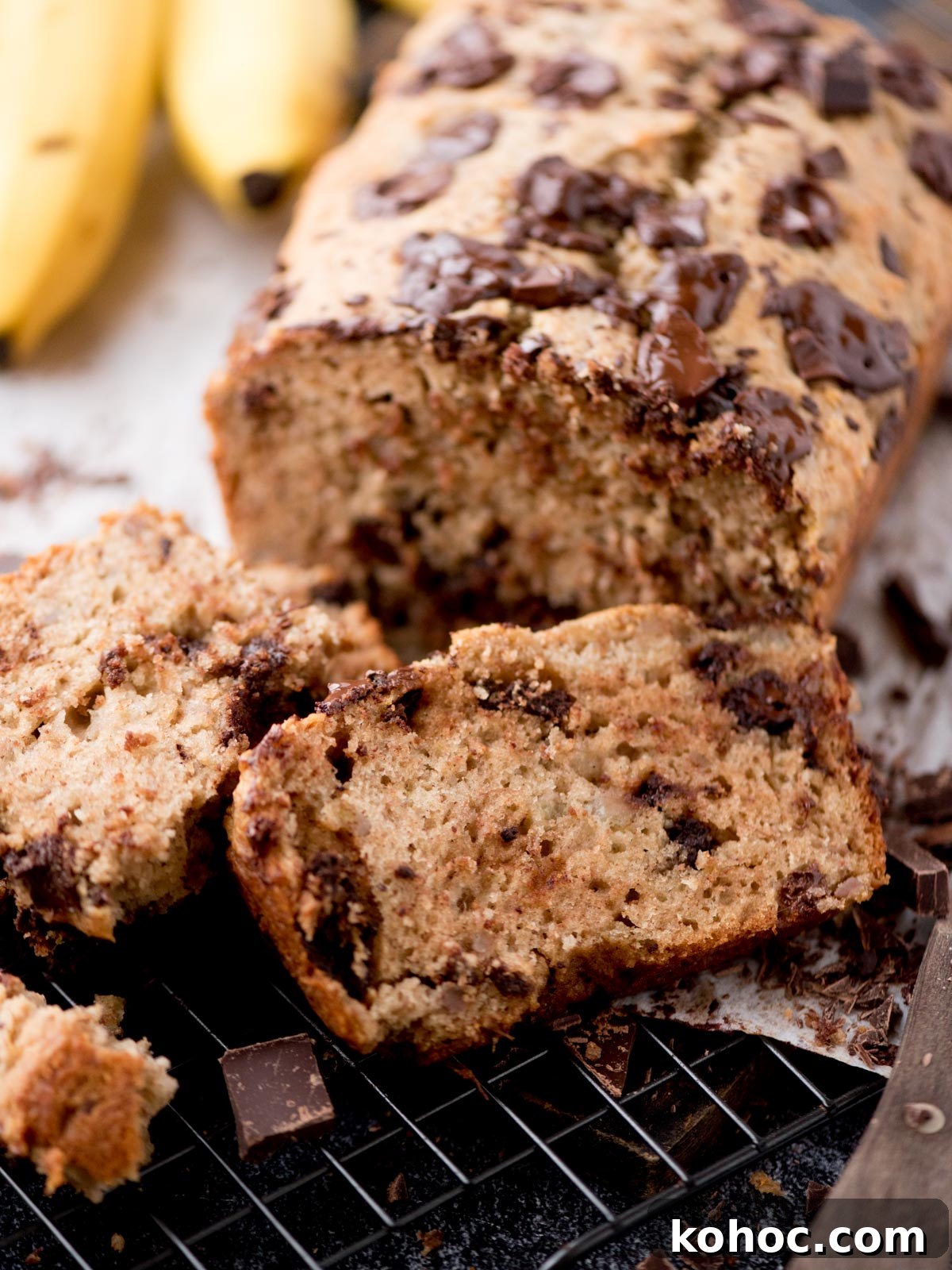 Chocolate Chunk Banana Bread Recipe with Melting Chocolate Chunks 10 A close-up of sliced chocolate chip banana bread on a cooling rack, with a loaf in the background and bananas partially visible. The bread is moist, dotted with chocolate chunks.